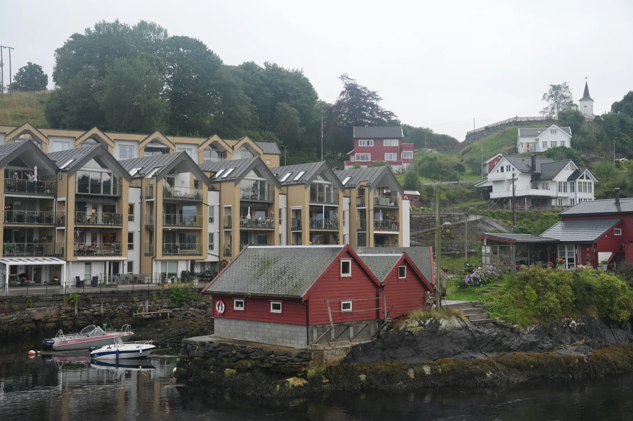 Rocky harbor with red boathouse, moored boats, apartment buildings, hillside houses, and a church steeple.