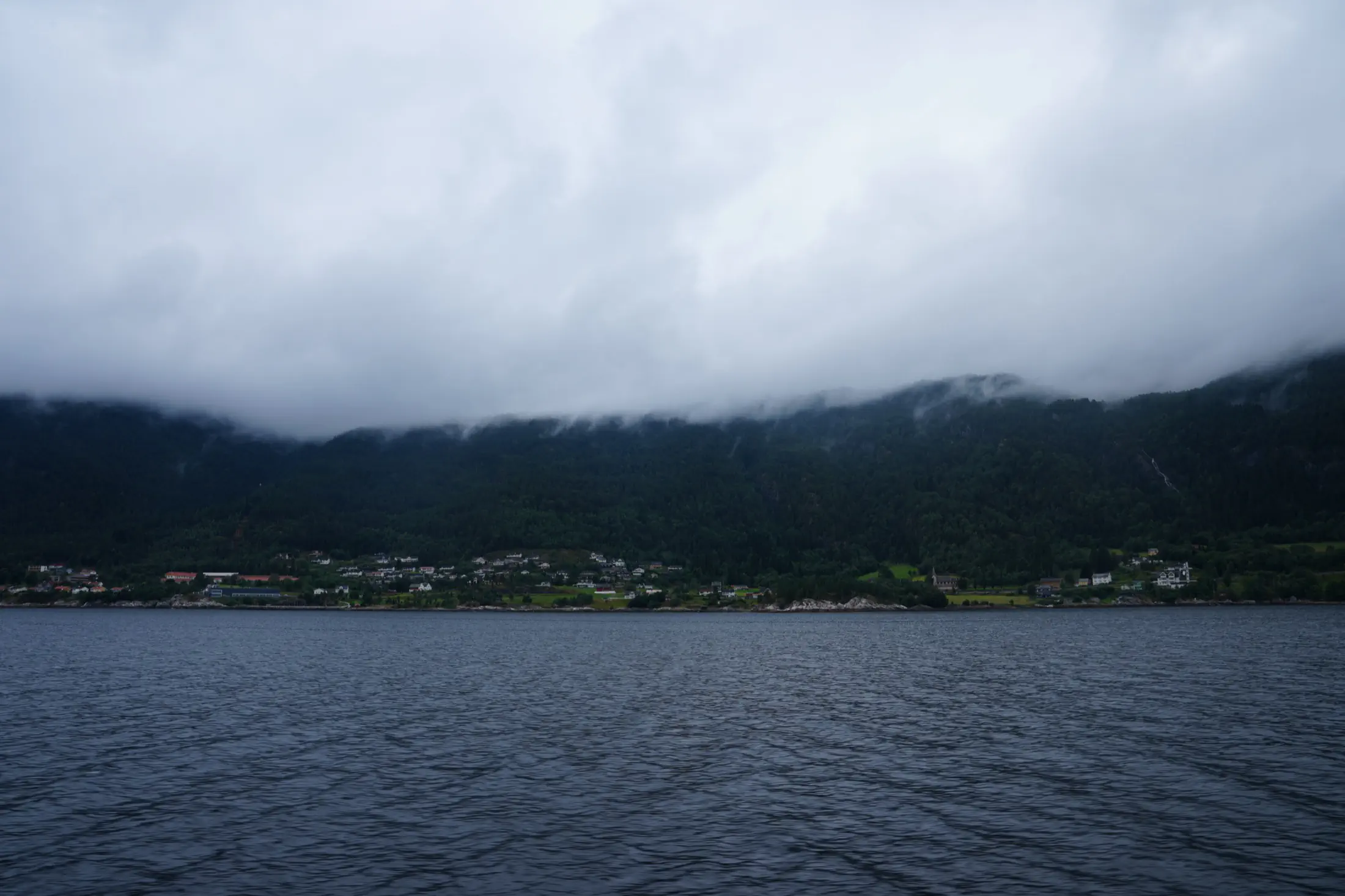 Calm water in the foreground, a small поселlement along the shore, and cloud-covered forested mountains behind it.