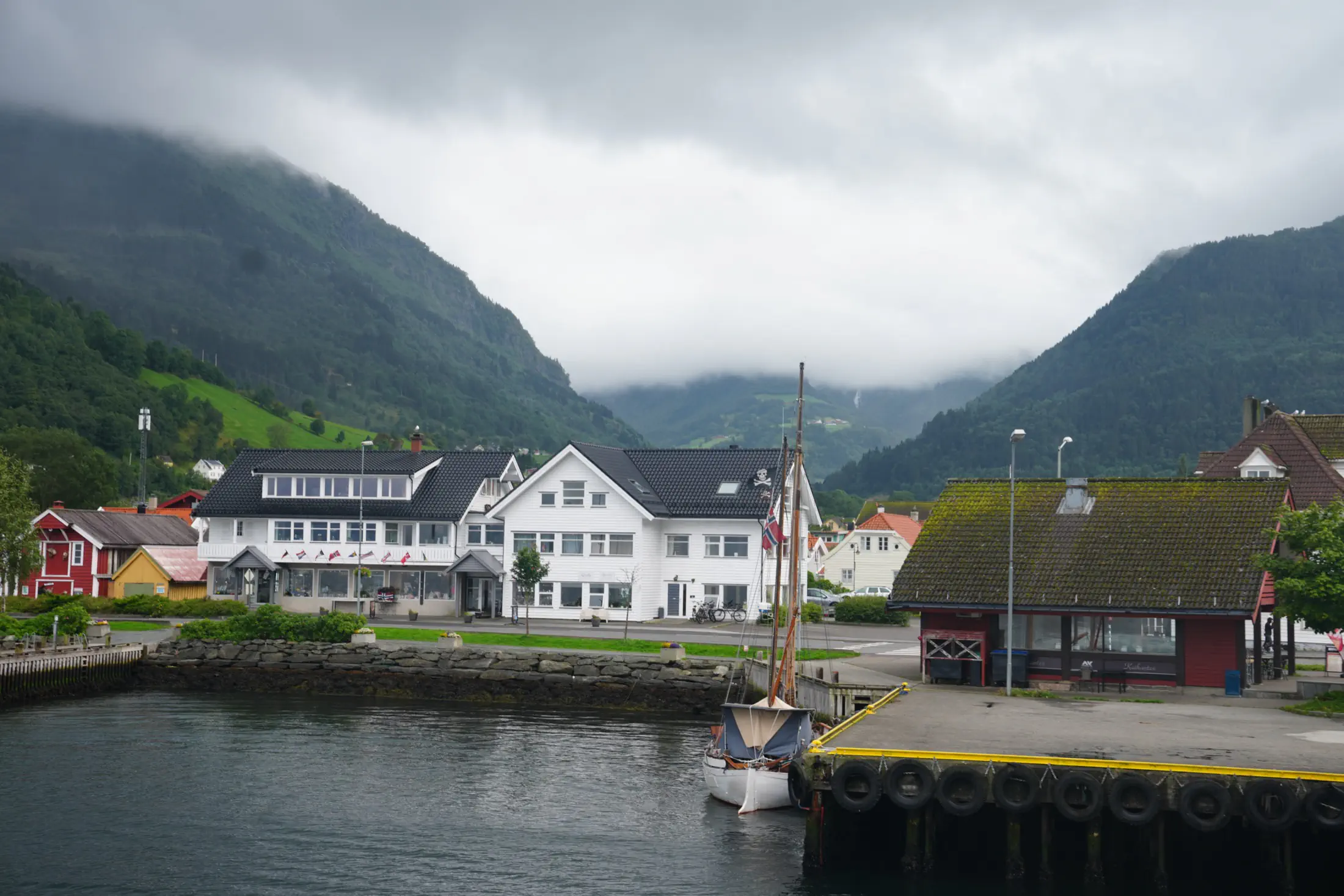 A small harbor with a moored sailboat, waterfront buildings, and steep mountains covered by low clouds.