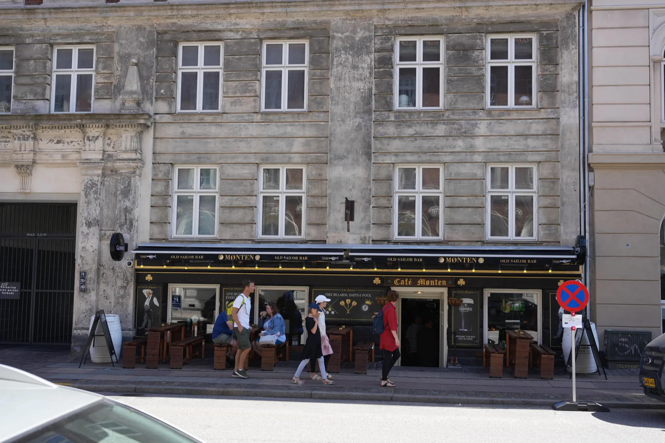 People standing and sitting outside Café Monten with wooden benches and outdoor tables along a city street.