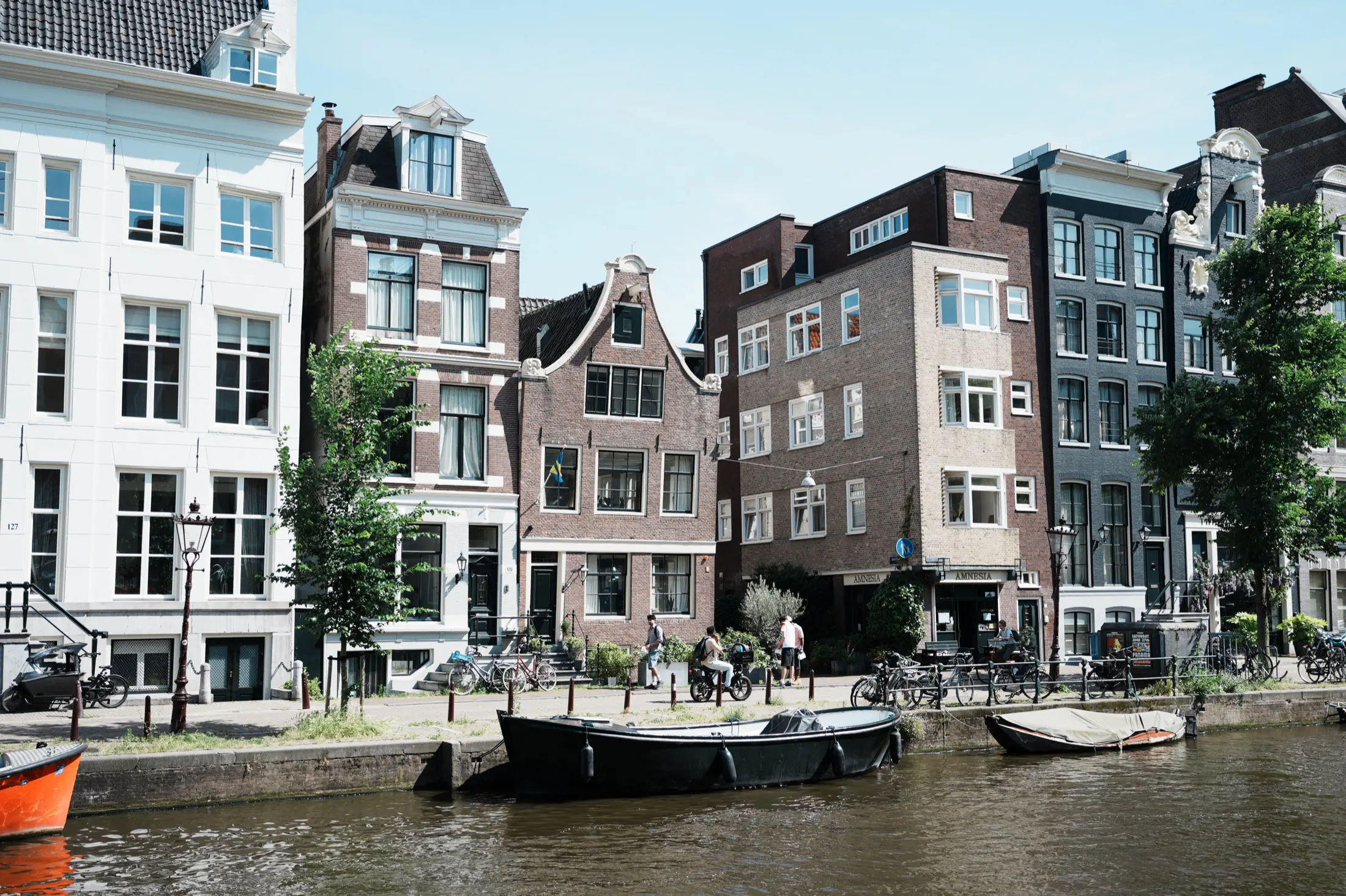Canal-side street in Amsterdam with historic buildings, bicycles, pedestrians, and boats moored along the water.